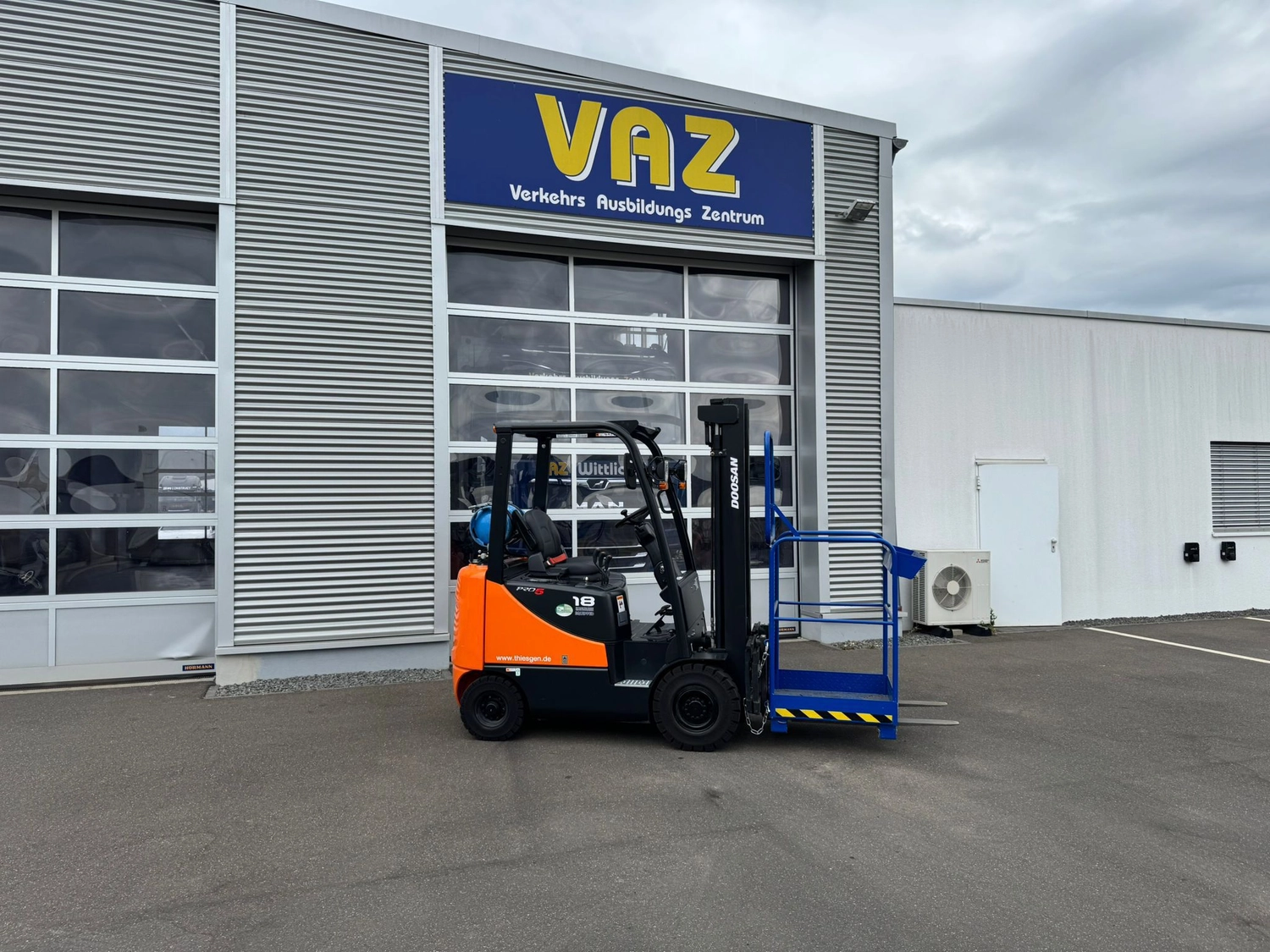 Medisem - Medizinische Seminare - Ein orange-schwarzer Doosan-Gabelstapler mit einem blauen Sicherheitskäfig an den Gabeln steht vor einem Gebäude mit der Aufschrift „VAZ Verkehrs Ausbildungszentrum“.