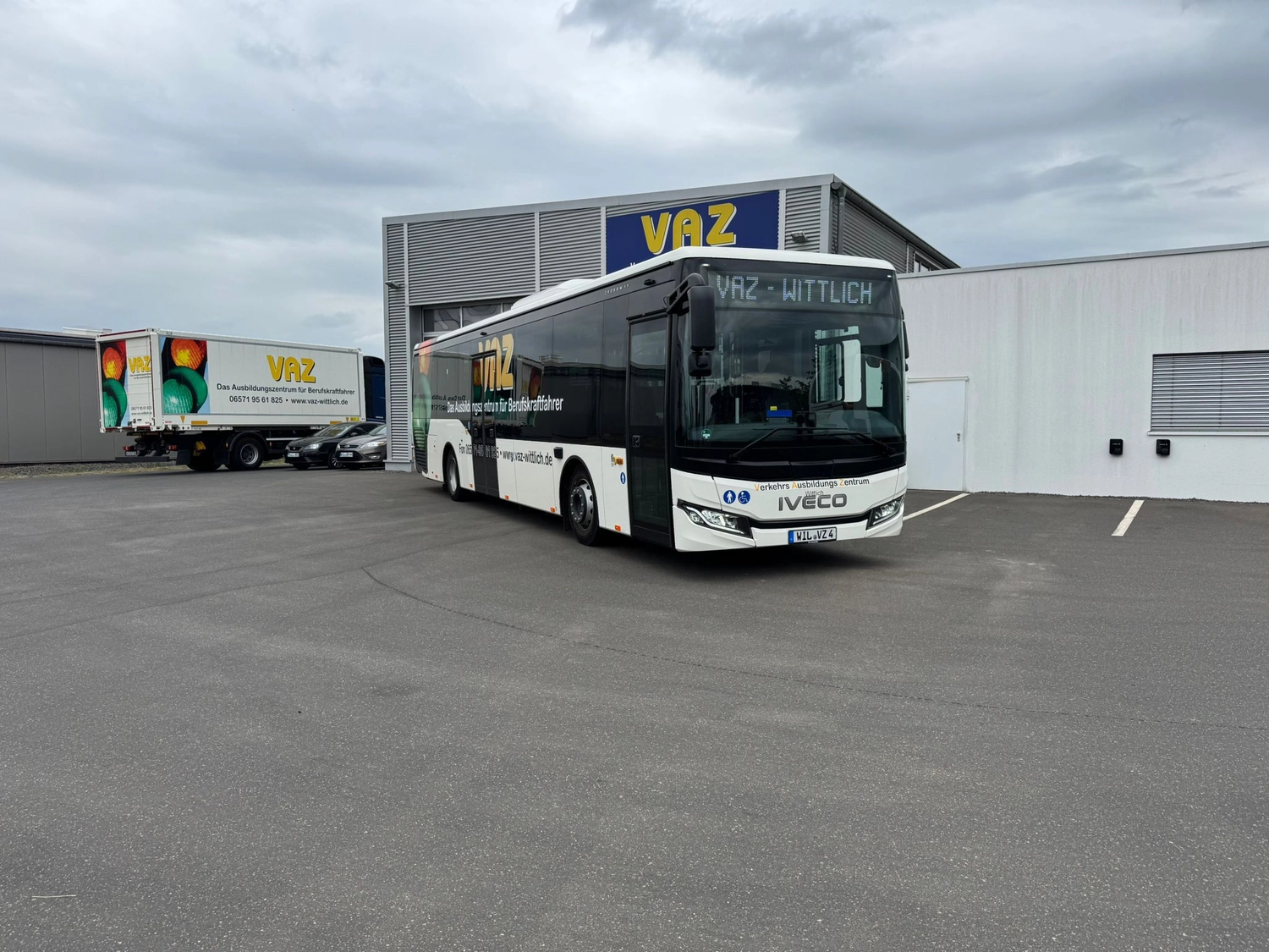 Medisem - Medizinische Seminare - Ein Iveco-Reisebus und ein Lastwagen parken vor einem VAZ-Industriegebäude unter bewölktem Himmel.
