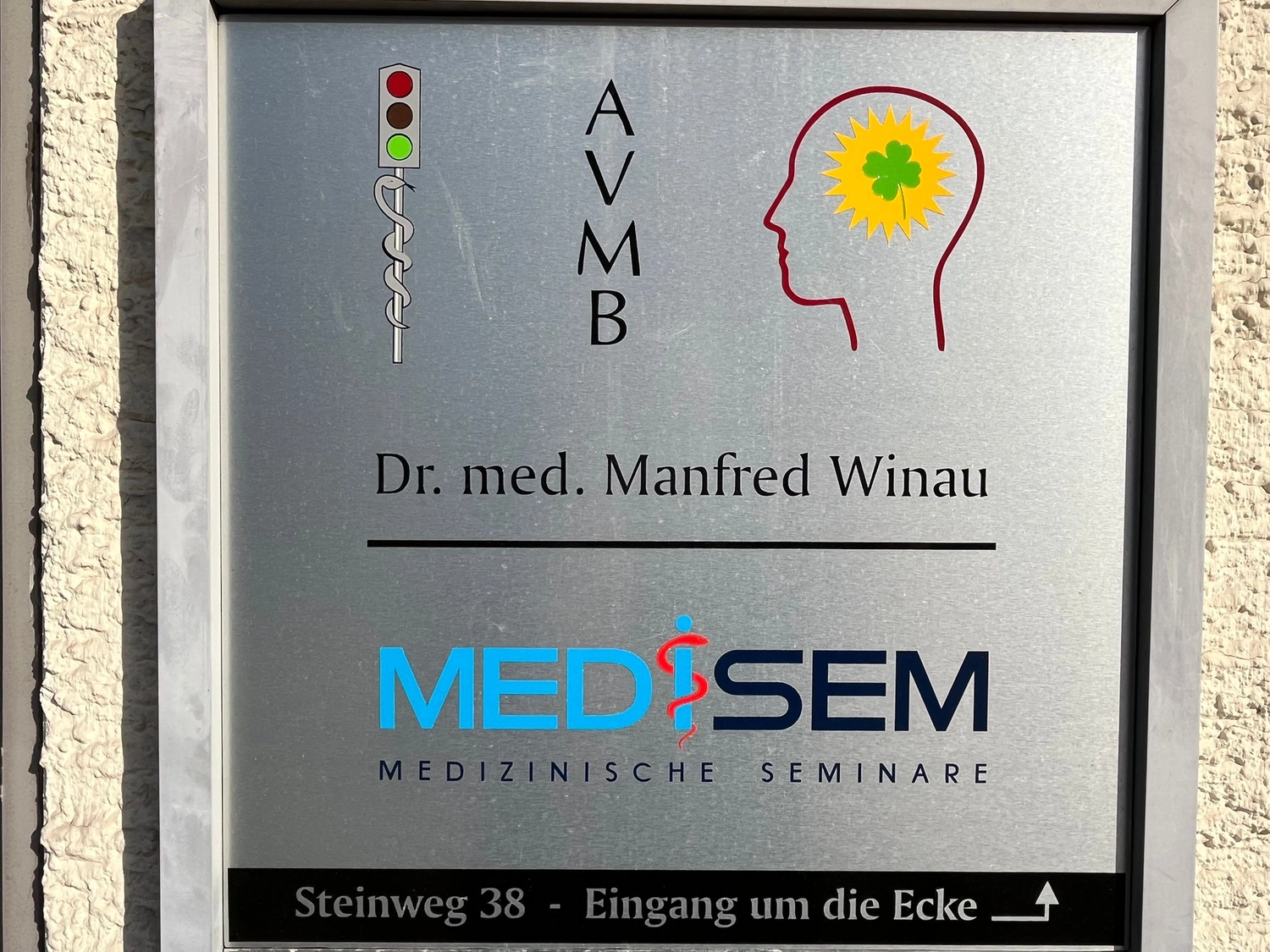 Medisem - Medizinische Seminare - Ein Metallschild an einem Gebäude mit der Aufschrift „Dr. med. Manfred Winau“ und „MEDISEM MEDIZINISCHES SEMINARE“ zeigt Logos einer medizinischen Ampel, die Silhouette eines menschlichen Kopfes mit gelber Sonne und die Adresse „Steinweg 38“.